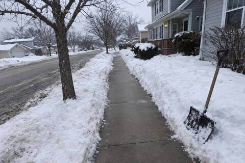 Public sidewalk cleared to bare concrete past an older two-story house, a shovel leaning on the snowbank beside it