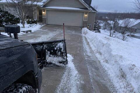 Pickup plow blade partway down a residential driveway that slopes toward an attached garage, snow curled off the blade edge
