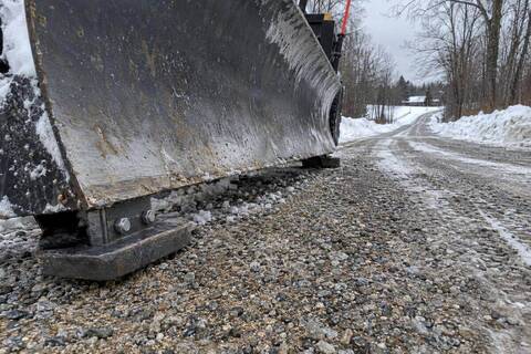 Close view of a plow blade held above a gravel driveway by bolted skid shoes, loose stone mixed through the plowed snow