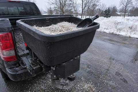 Open tailgate spreader hopper half full of coarse rock salt, a scoop resting on the hopper rim