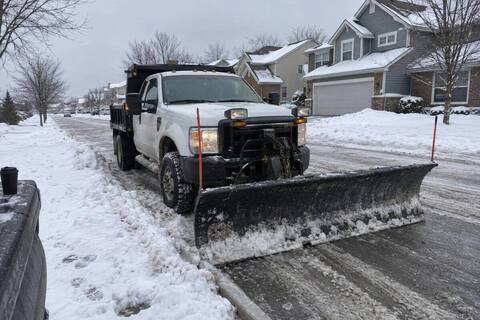 Plow truck parked at a residential curb between passes, blade lowered flat onto the snow-covered pavement