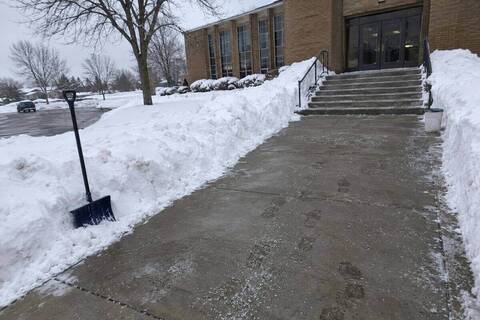 Wide concrete walkway cleared to a brick building's front steps, snow pushed back in high banks and grit on the treads