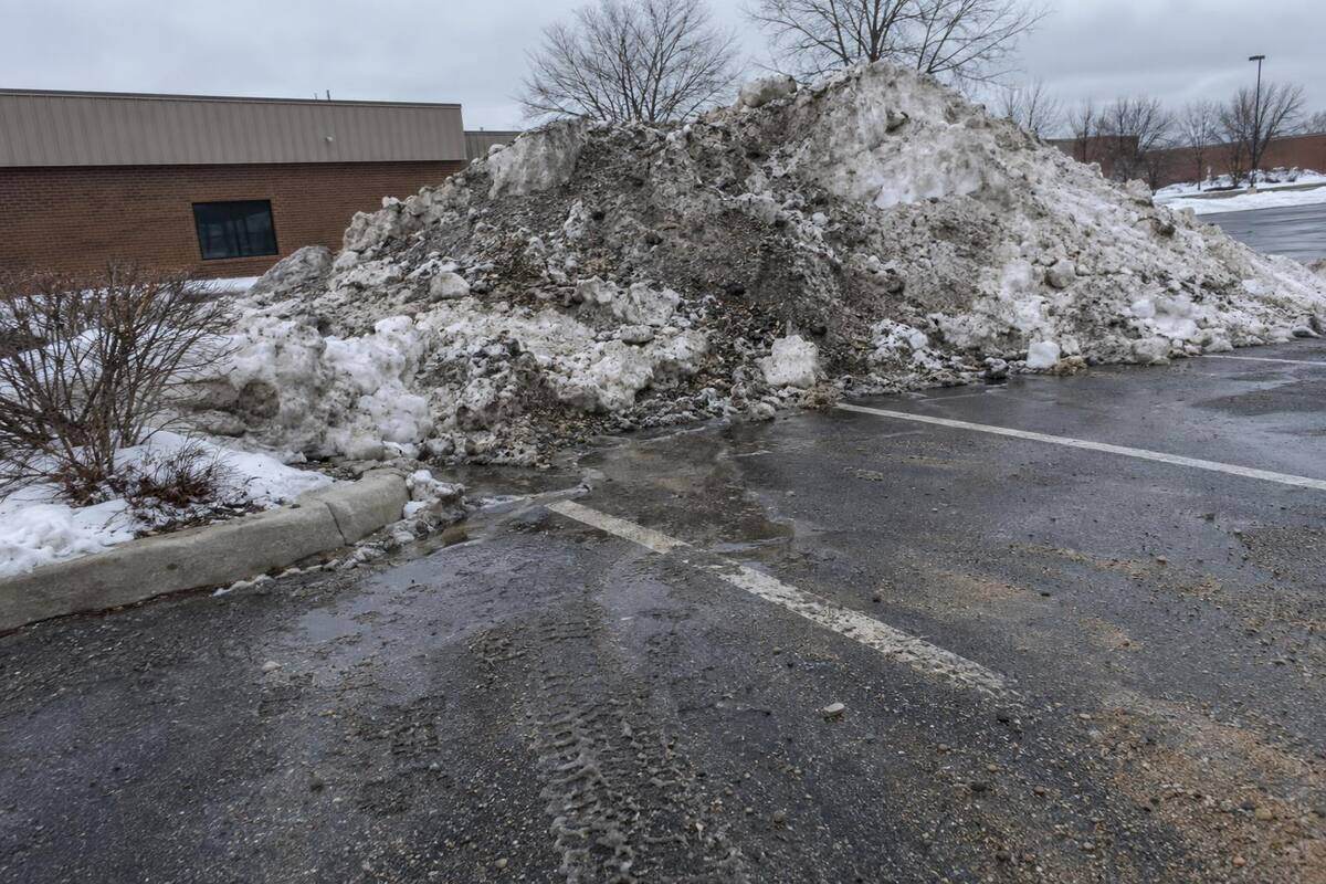 Pile of dirty compacted snow heaped at the edge of a commercial parking lot, meltwater running from its base
