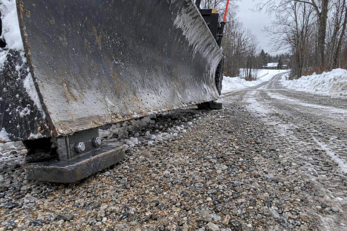 Close view of a plow blade held above a gravel driveway by bolted skid shoes, loose stone mixed through the plowed snow