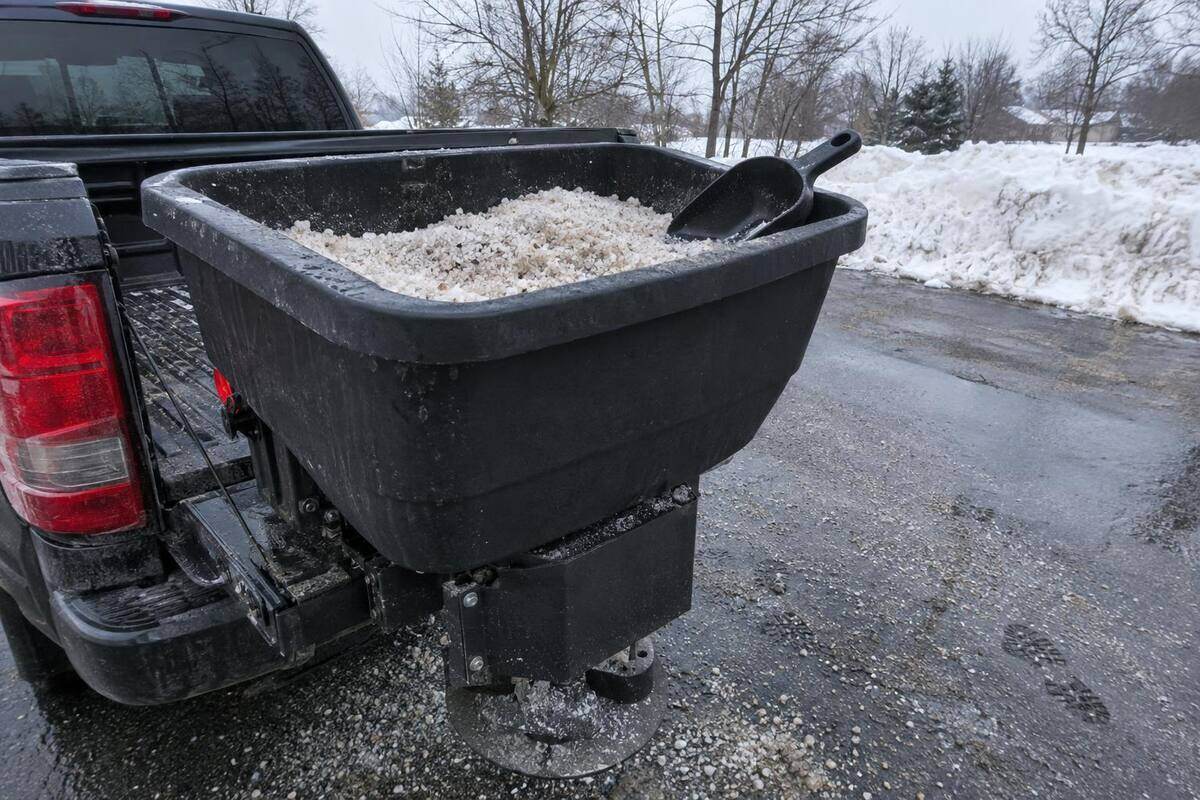 Open tailgate spreader hopper half full of coarse rock salt, a scoop resting on the hopper rim